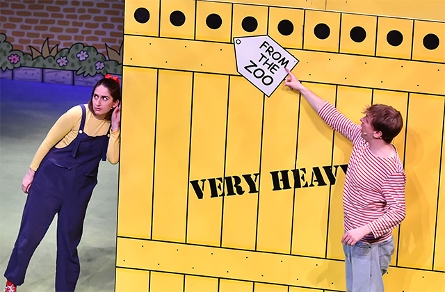 Two performers beside a huge yellow cartoonish crate labelled VERY HEAVY.