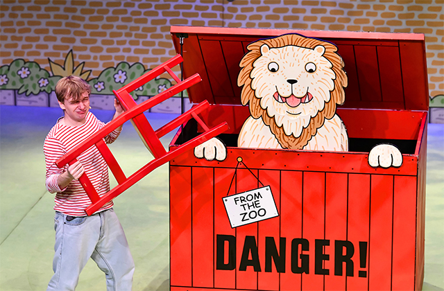 A performer attempting to put a red chair in a large red crate labelled DANGER with a cartoon lion inside.