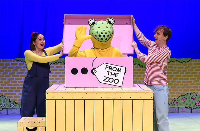 Two performers opening a pink box with a large, costumed frog inside.