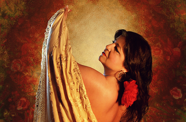 A woman in profile holds up a draped golden shawl, her bare shoulders lit warmly against a rich, textured red and orange backdrop. She wears large hoop earrings and a vivid red flower in her dark hair, evoking a flamenco-inspired, dramatic pose.