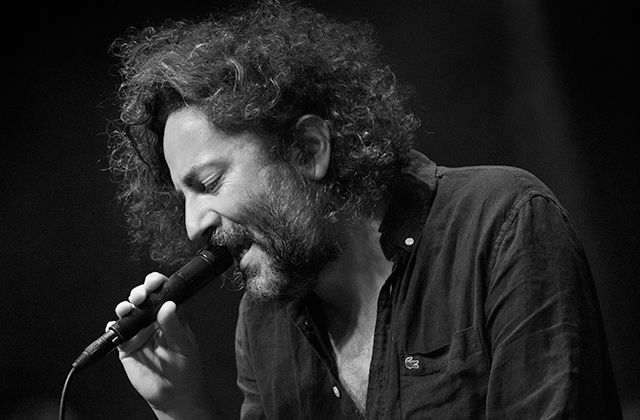 Black-and-white photograph of a person performing on stage, shown in close-up profile while holding a handheld microphone near the mouth. The person has curly hair and facial hair and wears a dark, buttoned shirt with an open collar. The microphone cable runs downward out of frame. Soft stage lighting illuminates the face and upper body, while the background fades into deep shadow, emphasizing the performer and the microphone in the foreground.