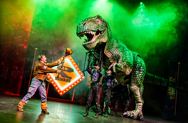 Performer on stage holds a glowing “Beware” dinosaur warning sign while a giant T-Rex puppet towers overhead in green theatrical lighting and smoke. Several puppeteers dressed in dark costumes control the dinosaur during the live stage show.