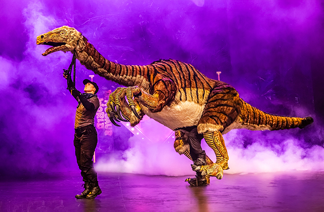 A large feathered dinosaur puppet with striped orange and brown markings stretches across the stage under purple lighting and fog, operated by two visible performers in black costumes.