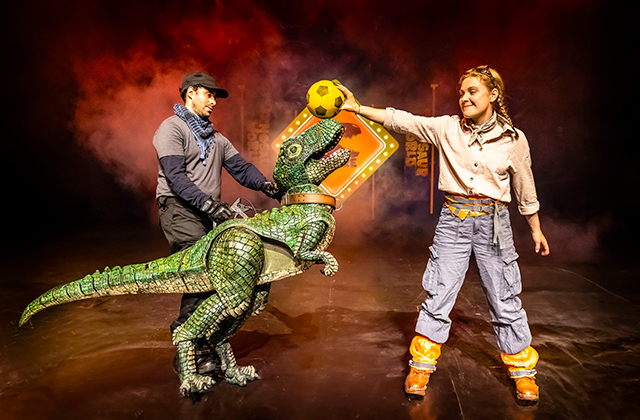 A performer smiles while holding a football above a small green dinosaur puppet’s head on stage. Another puppeteer supports the dinosaur from behind, with red lighting and smoke effects in the background.