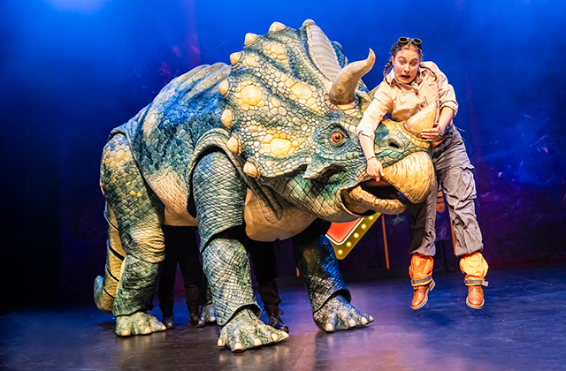 A large blue triceratops-style dinosaur puppet stands on stage while a performer appears lifted or tugged by one of its horns. Blue theatrical lighting illuminates the scene.