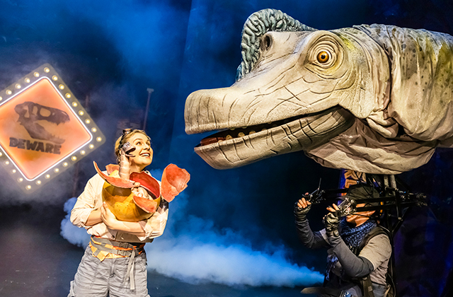 A smiling performer holds a dinosaur egg and shell pieces while facing a large long-snouted dinosaur puppet operated by a visible puppeteer. A glowing “Beware” sign and stage smoke appear in the background.