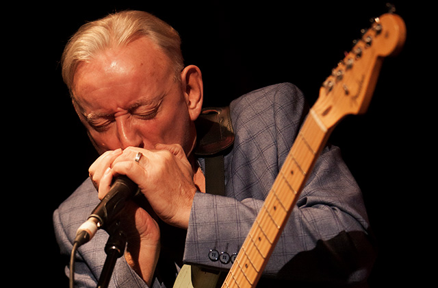 Don Baker playing the harmonica, with a guitar hanging in front of him