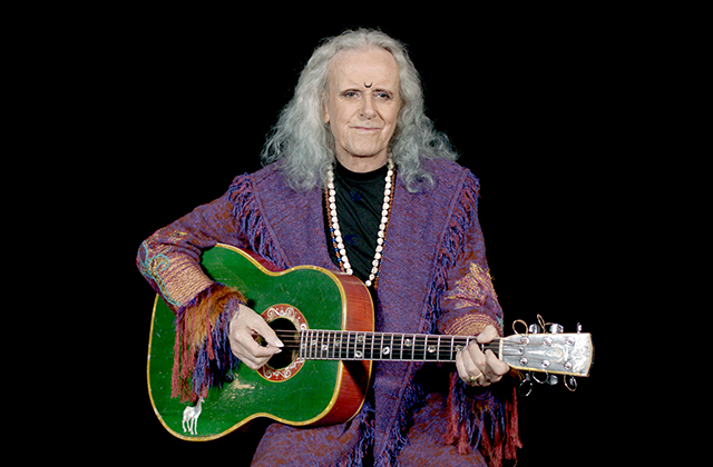 Donovan with long, wavy grey hair sits against a black background holding a green acoustic guitar. He is wearing a purple fringed jacket over a black top, a long beaded necklace, and a small crescent moon symbol on his forehead.