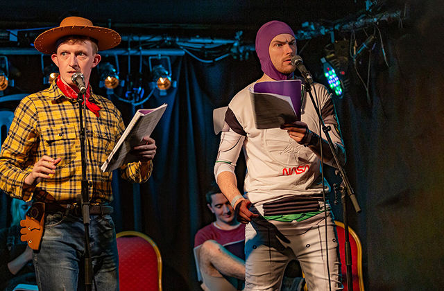 Stephen Colfer, dressed as Woody from Toy Story, and Gavin Drea, dressed as Buzz Lightyear, speaking into microphones on a stage.