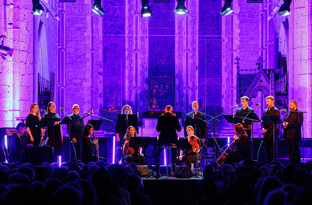 A large ensemble of instrumentalists and singers assembled in front of a conductor with a dramatic purple backdrop.