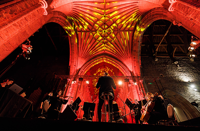 A dramatic upward-facing shot of a conductor leading a large ensemble with an arched church ceiling above.
