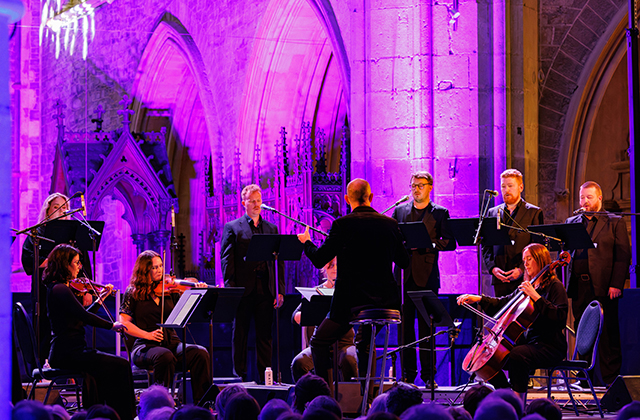 Mid-shot of a conductor leading an ensemble of singers and instrumentalists against a purple backdrop.