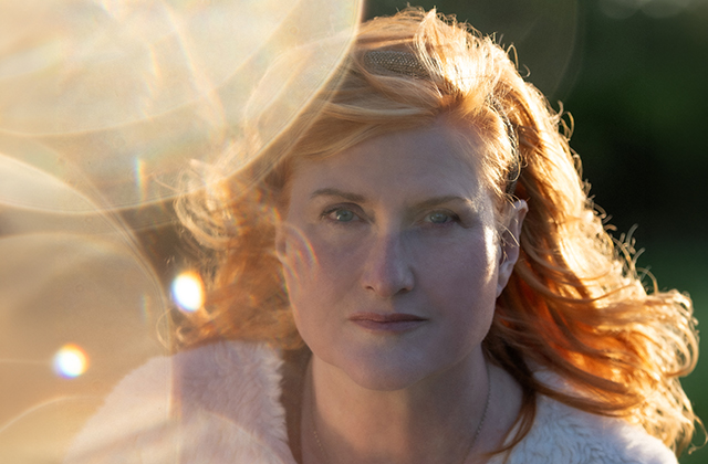 Eddi Reader is pictured looking upward towards the camera, she is wearing a white top. The image is very light with some atmospheric lens flare and warm colour.