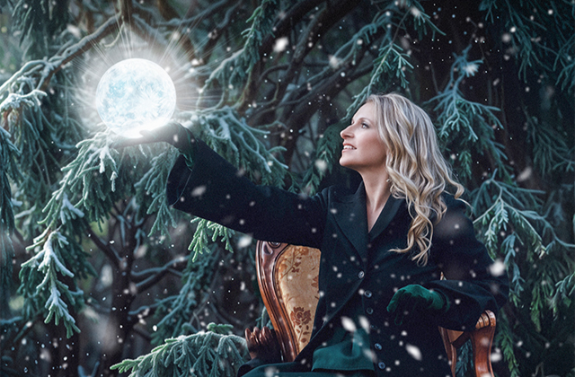 Eimear Quinn sits on an ornate chair in a snowy forest, surrounded by frosted evergreen branches. She is wearing a dark coat and green gloves, smiling as she holds a glowing orb resembling the moon in one outstretched hand. Snowflakes gently fall around her, adding to the magical, wintery atmosphere.