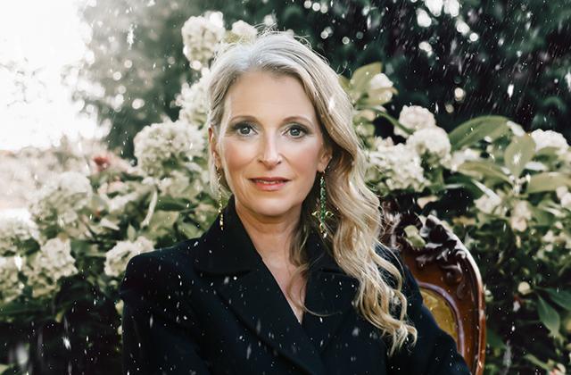 Portrait of Eimear Quinn outdoors, wearing a black blazer and green earrings, seated in front of blooming white flowers with soft snow falling around her