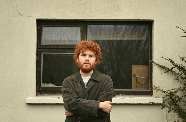 Colour photograph of a person standing outdoors in front of a cream-coloured wall with a single rectangular window. The person stands centered in the frame with arms crossed, wearing a dark jacket over a light top. The window behind has dark trim and reflects bare tree branches, with a small cardboard box visible on the interior windowsill. Sparse greenery climbs the wall on the right edge of the image, and soft natural light gives the scene a muted, earthy tone.