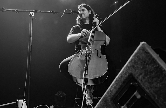 Black-and-white photo of a musician seated on a stage, playing a cello. The cello rests between the performer’s knees, with one hand on the fingerboard and the other near the bow. A microphone on a stand is positioned close to the performer, and stage lights illuminate the scene from above. Audio cables, a microphone stand, and stage equipment are visible in the foreground and background, suggesting a live performance setting.