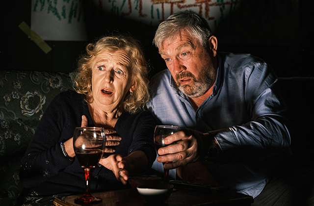 Joan Sheehy and Seamus O'Rourke, with glasses of red wine, are lit with the glow of a television, and looking shocked