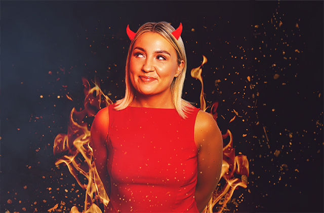 Enya Martin, a smiling woman with blonde hair stands in front of stylized flames. She is wearing a bright red sleeveless dress and small red devil horns. She looks playfully upward with a mischievous expression. The background is dark, with glowing sparks and fiery effects surrounding her.