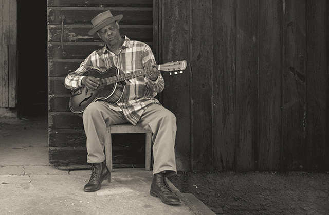 A black and white image of Eric Bibb sitting with his guitar wearing a checked shirt, in a typical