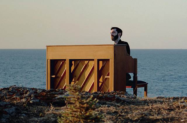 Evgeny Grinko sits at a wooden upright piano with a still coastal background