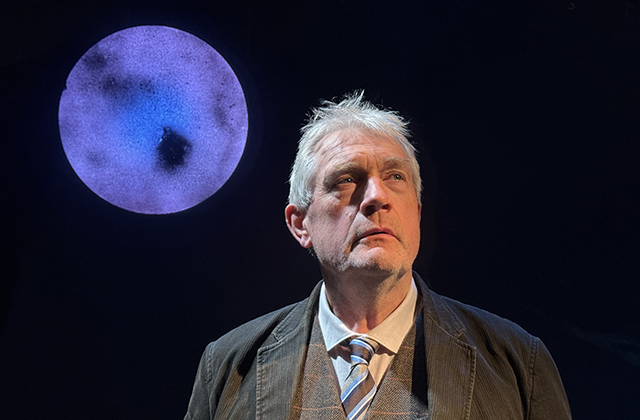 Seamus O'Rourke, dressed in a grey suit, stands in front of a blue full moon