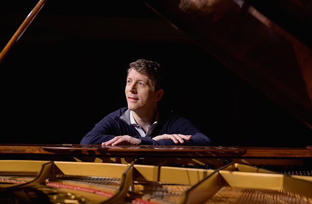 Finghin Collins sits at an open grand piano, casually dressed and looking off the left, with his arms resting on the top