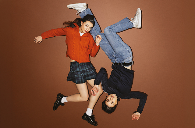 Two young women are captured mid-air against a plain brown background, creating a playful, gravity-defying effect. One, in a red cardigan and plaid skirt, appears upright, while the other, in a dark top and jeans, is upside down.