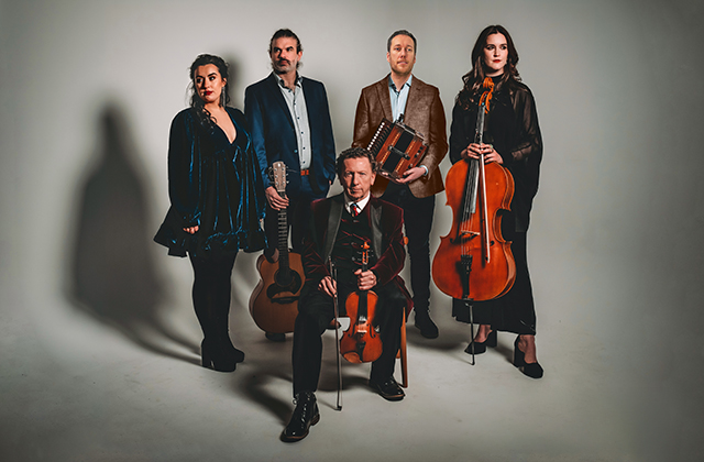 A promotional studio portrait of a five-member traditional Irish music ensemble against a light grey background. In the center, traditional musician Frankie Gavin sits on a chair wearing a dark velvet jacket and holding a fiddle and bow. He is surrounded by four standing musicians: a woman in a short dark velvet dress on the left, a man holding an acoustic guitar, a man holding an accordion, and a woman holding a cello on the right.