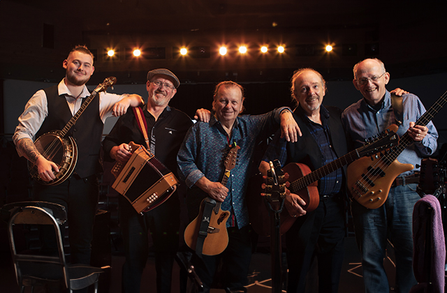 A photo of the five members of The Fureys, each holding their respective instrument (banjo, accordion, two with acoustic guitar, and bass guitar), smiling and leaning on one another, in what appears to be a music venue or theatre.