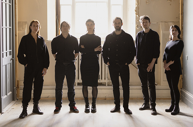 Six members of Crash Ensemble wearing all black, in a sparse room with their backs to a bright window.