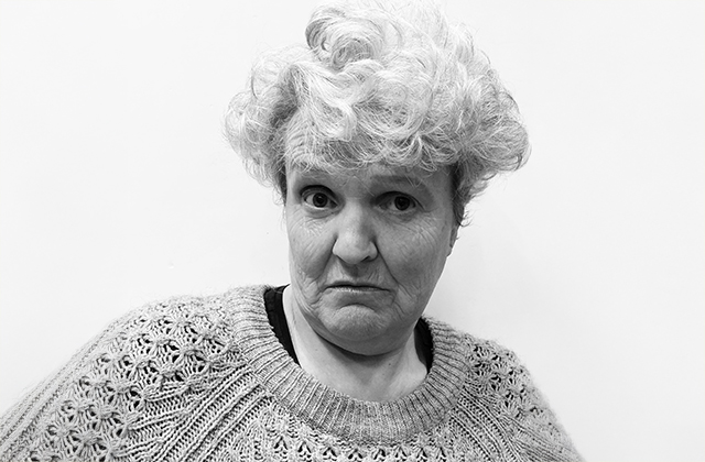 Rose Henderson wearing a knitted jumper, staring quizzically down the camera lens. The image is in black and white.