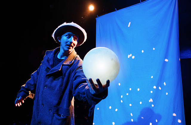 The actor from Hay! stands on stage lit up by a strong blue colour, he is holding out a glowing orb which looks very moon like.