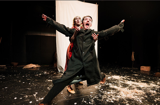 The two actors from Hay! are pictured centre stage. The man stands with his arms out, his leg is sliding out in front of him towards the camera, he has a clumsy look on his face. The woman stands behind holding underneath his arms in an attempt to catch him