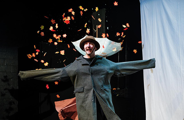 Actor from Hay! He stands centre shot, wearing a hat and a lock black coat, there is an umbrella falling upside down behind him, his arms are straight out either side and lots of autumn leaves are falling in the air.