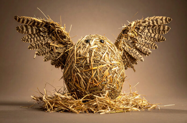 A photograph of an owl sitting in an nest, both the nest and the owl are made out of hay, the owl has its wings spread up and wide.
