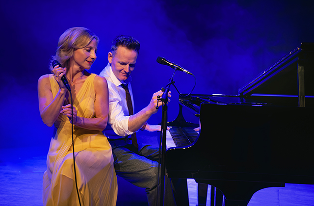 A female singer in a flowing yellow dress stands beside a grand piano, holding a microphone and smiling toward a male pianist seated at the keys. Blue stage lighting and light haze create an intimate concert atmosphere.