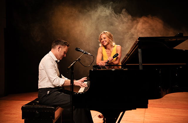 On a warmly lit stage, a male pianist plays a grand piano while a female singer in a yellow dress leans on the instrument, smiling and holding a microphone. Soft amber lighting and light smoke add a cosy, theatrical mood.