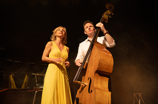 A female singer in a yellow dress looks upward while a male musician beside her plays a double bass. The stage is warmly lit with a smoky backdrop, suggesting a live jazz or cabaret-style performance.