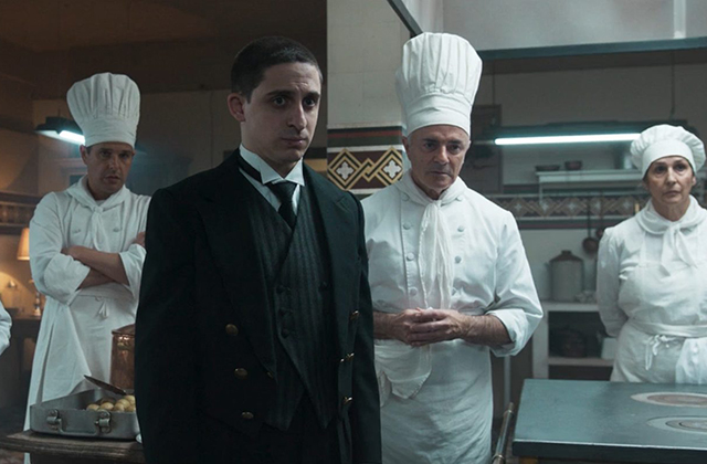 A man in a formal black suit, waistcoat, and white-collared shirt stands prominently in the foreground of a professional kitchen with a serious expression. Surrounding him are three kitchen staff members dressed in traditional white chef jackets and tall white toque hats: a man with arms crossed to the left, a man standing central with hands clasped in front of him, and a woman standing to the right.