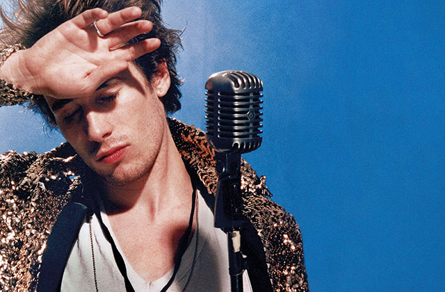 A close up of Jeff Buckley, he stands in front of a microphone with a his hand across his forehead and his eyes closed looking off to the distance. He's wearing a white t-shirt, snake skin pattern jacket against a blue background.