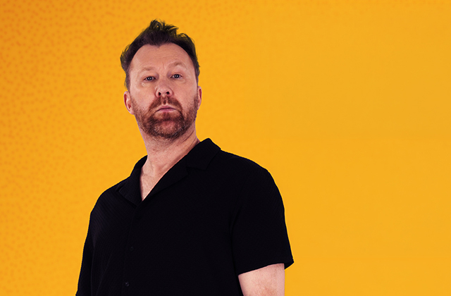 Jason Byrne stands staring towards the camera with a firm expression. He is wearing a black short sleeve shirt against an orange background.