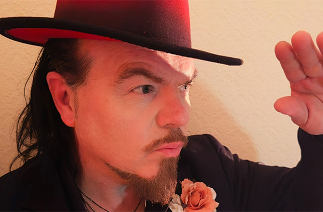 Jack L wearing a wide-brimmed hat with a red underside looks to the side, raising his hand slightly as if shading his eyes. He has a short beard and moustache, long hair, and wears a dark jacket with a pink rose on the lapel.