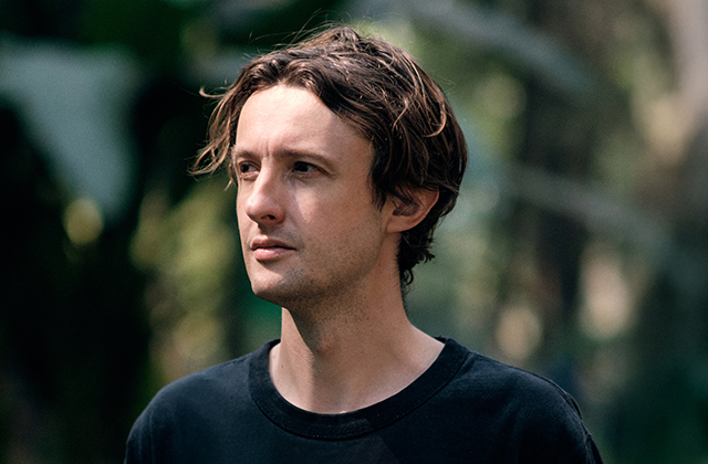 A close-up portrait of a light-skinned man with wavy brown hair looking off-camera to the left. He is wearing a plain black t-shirt against a blurred green, natural foliage background.