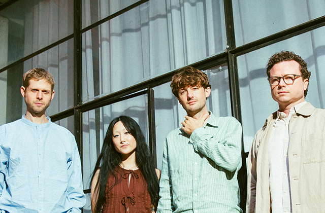 A outdoor group portrait of four people standing side-by-side in front of a modern glass-paneled building with sheer white curtains visible inside. From left to right: a man in a light blue collarless shirt, a woman with long dark hair in a rust-brown sleeveless top, a man in a green striped linen button-up holding his hand near his collar, and a man with glasses wearing a beige jacket over a light shirt.