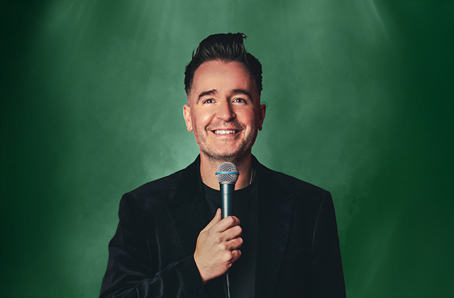 Jarlath Regan in a black suit stands holding a microphone up to his smiling face, with a green backdrop