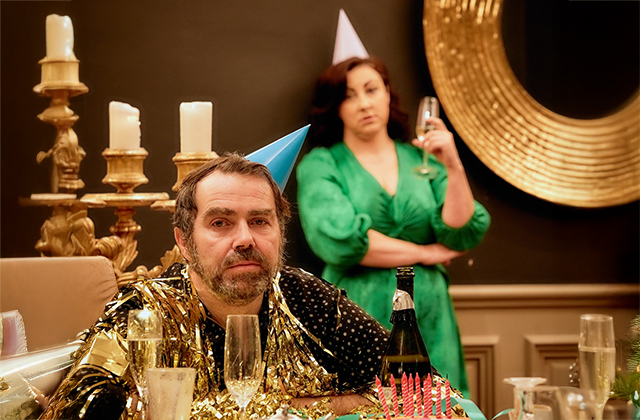 Pat Ryan sits at a cluttered party table wearing a shiny gold foil garland and blue party hat, looking dishevelled and weary. Behind him, Georgina Miller stands in a bright green dress, holding a champagne glass with folded arms and a displeased expression. The atmosphere is tense despite the festive decorations.
