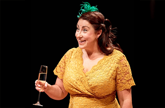 Georgina Miller beams with a raised eyebrow and a delighted expression, holding a champagne flute. She wears a yellow lace dress with a green feathered fascinator, suggesting a festive or celebratory moment in the play.