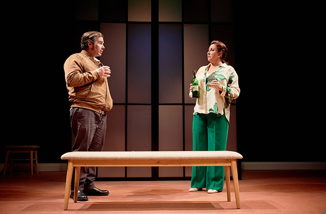 Pat Ryan and Georgina Miller face each other across a wooden bench in a minimalist set. He holds a glass, while she clutches two bottles. Their expressions are serious, suggesting a moment of tension or confrontation.