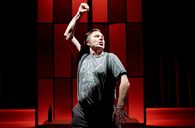 Pat Ryan, in a graphic t-shirt and suspenders, stands alone under dramatic red lighting with one fist raised in the air. His expression is defiant and intense, capturing a dynamic and emotionally charged moment.